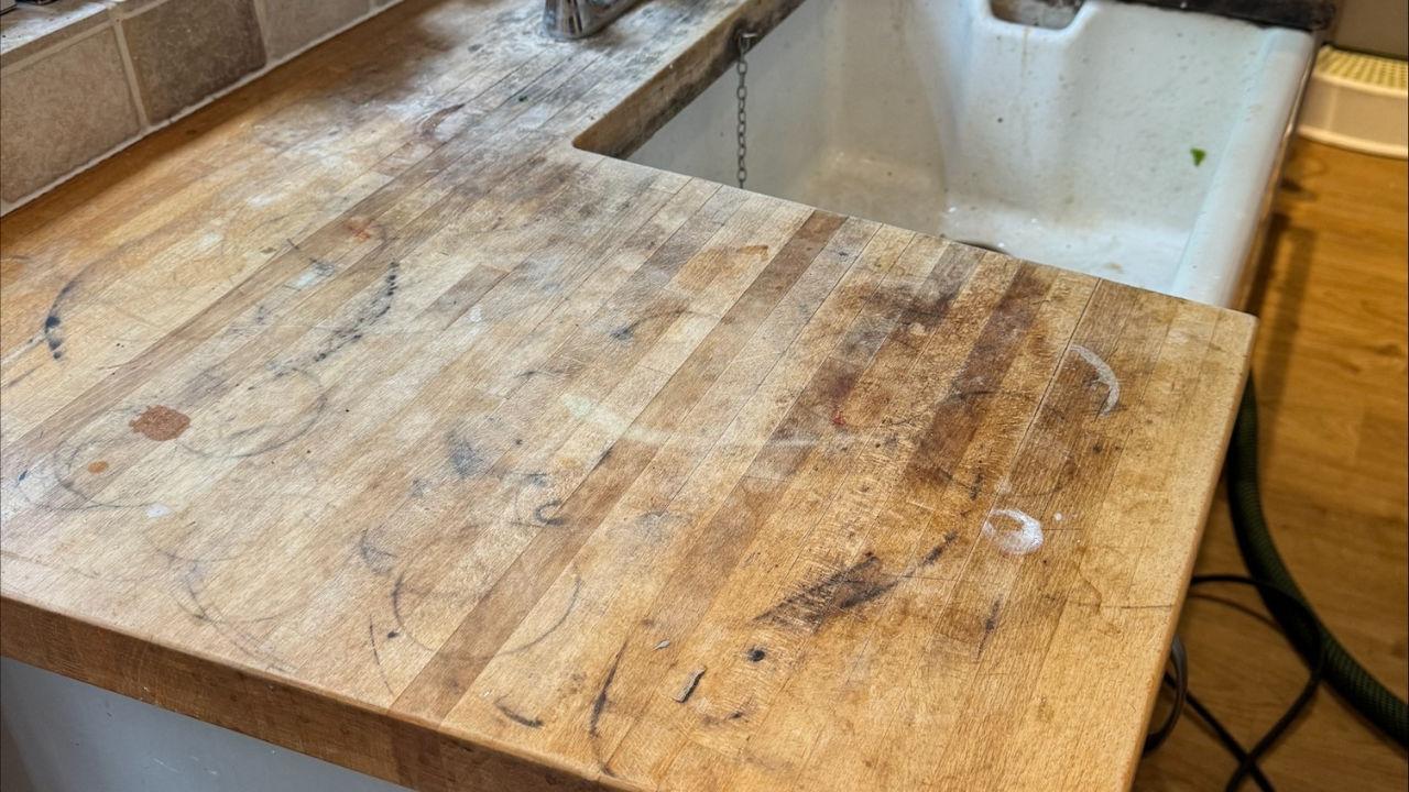 Heavily worn oak butcher-block kitchen worktop before Worktop Refinishing in southwoodford, beside a white ceramic sink and engineered oak floor, showing stains, rings and scratches.